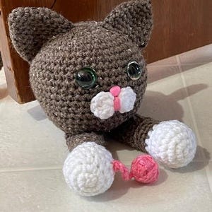 May include: A crocheted brown cat with white paws and a pink tail. The cat has large green eyes and a pink nose.