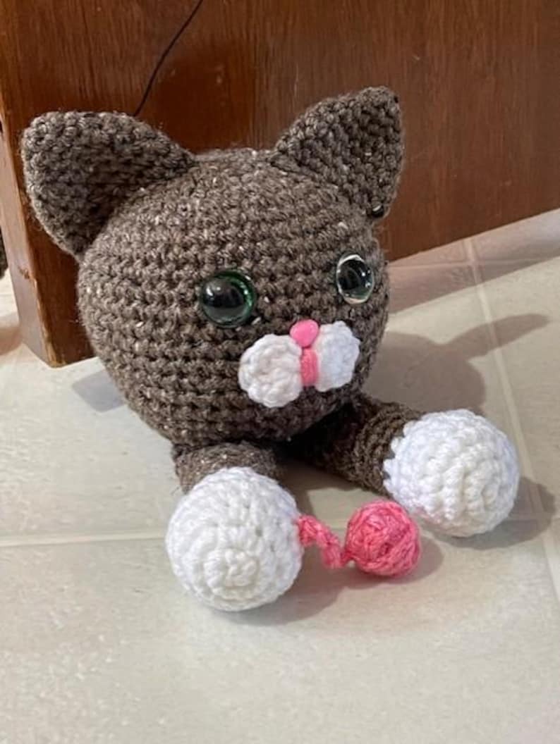 May include: A crocheted brown cat with white paws and a pink tail. The cat has large green eyes and a pink nose.