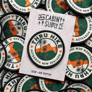 Op de afbeelding: Ronde opstrijkbare patch met de tekst "THRU HIKE AOTEAROA NEW ZEALAND" rond een landschapsontwerp in groen, oranje en wit. De patch zit op een kaart met "the CABIN SUPPLY CO" erop gedrukt. Andere patches staan op de achtergrond.