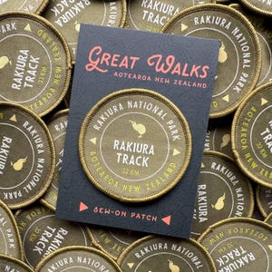 Könnte beinhalten: Runder, olivgrüner Aufnäher mit der Aufschrift "Rakiura Track 32 KM" und einer Kiwi-Vogel-Illustration. Teil der "Great Walks Aotearoa New Zealand" Kollektion. Die Aufnäher haben einen goldenen Rand.