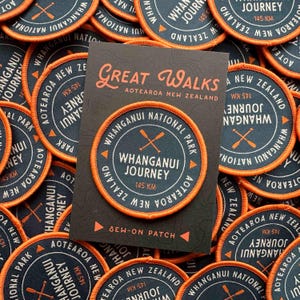 May include: Circular sew-on patches with an orange border and navy blue center. The patches feature the text "Whanganui Journey 145 KM" and "Whanganui National Park, Aotearoa New Zealand". A card with the text "Great Walks Aotearoa New Zealand" is in the center.