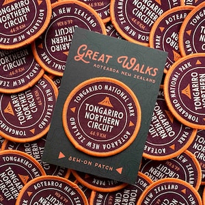 Puede incluir: Un conjunto de parches redondos para coser y una tarjeta. Los parches son de color burdeos con bordes naranjas, con el texto "Tongariro Northern Circuit 44,9 km" y "Aotearoa New Zealand". La tarjeta dice "Great Walks Aotearoa New Zealand".