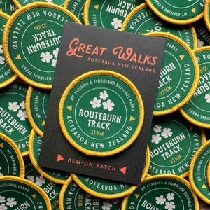 Può includere: Toppe ricamate verdi e gialle con la scritta "ROUTEBURN TRACK 33 KM AOTEAROA NEW ZEALAND" e "Great Walks Aotearoa New Zealand". Le toppe presentano disegni di fiori bianchi e sono da cucire.