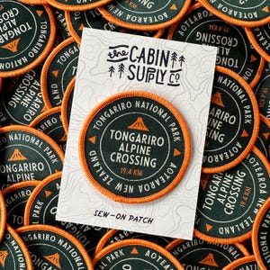 May include: Circular sew-on patches with an orange border and dark green center. The patches feature the text "Tongariro Alpine Crossing" and "19.4 KM" with a mountain graphic. The patches are from "The Cabin Supply Co."