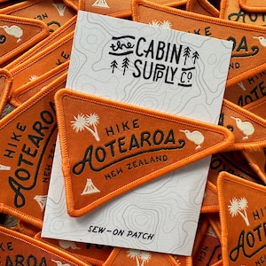 Peut inclure: Écussons triangulaires orange à coudre avec le texte "Hike Aotearoa New Zealand" et une illustration d'un kiwi. Les écussons sont entourés d'autres écussons et d'une carte avec le texte "The Cabin Supply Co."