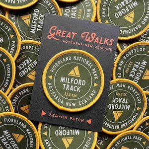 Puede incluir: Un conjunto de parches de color verde oliva y dorado con el texto "Milford Track" y "Fiordland National Park". Los parches rodean una tarjeta negra con las palabras "Great Walks Aotearoa New Zealand".