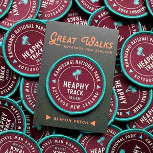 Könnte beinhalten: Eine Sammlung runder Aufnäher mit türkisfarbenem Rand und weinrotem Zentrum. Die Aufnäher tragen den Text "Heaphy Track 78,4 km" und "Kahurangi National Park, Aotearoa New Zealand". Eine Karte mit "Great Walks" ist ebenfalls sichtbar.