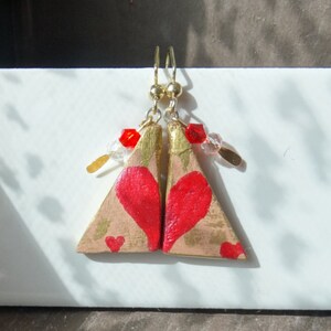 Può includere: Orecchini fatti a mano con un design triangolare, ognuno con un cuore rosso dipinto. Gli orecchini hanno dettagli dorati, ganci dorati e sono adornati con perline rosse e trasparenti. Un accessorio artistico unico.