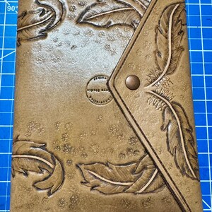 May include: A tan leather journal with embossed feather designs and a flap closure. The journal features a button clasp and the text "toys studio" on a circular emblem. The cover is decorated with multiple feather patterns.