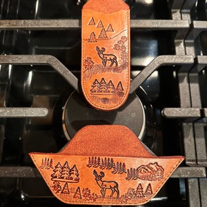 May include: Two brown leather knife sheaths with a nature-themed design. The sheaths feature a deer, trees, and mountain landscape. The top sheath is a vertical, elongated shape, while the bottom sheath has a wider, curved design.