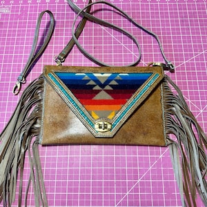 May include: Brown leather crossbody bag with a colorful geometric design on the flap. The bag features long fringe on the sides, a detachable strap, and a wristlet strap. The flap has a turquoise beaded trim and a gold-tone clasp.
