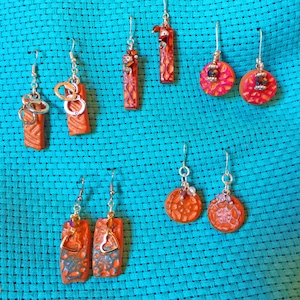 May include: A collection of handmade earrings with various designs. Some earrings feature rectangular orange blocks with silver handcuffs or heart charms. Other earrings have round orange and pink designs with decorative elements. The earrings hang against a turquoise background.