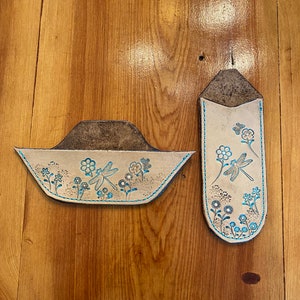 May include: Two leather items with dragonfly and floral designs. One is a small, open-topped container with a blue stitched border. The other is a tall, narrow case with a similar design. Both are tan with blue and brown accents.