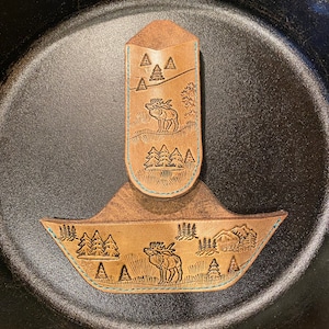 May include: Two brown leather pan handle covers with a nature scene featuring trees, a moose, and a cabin. The covers are stitched with turquoise thread. One cover is rectangular, and the other is a curved shape.