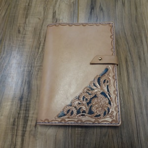 May include: Tan leather journal with a decorative corner design. The corner features a floral pattern with blue accents and a scalloped edge. A leather strap with a brass snap closure secures the journal.