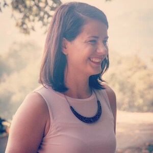 May include: A woman with short brown hair is smiling and wearing a light pink sleeveless top with a black geometric necklace.