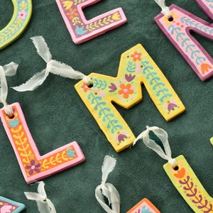 May include: A set of colorful, hand-painted wooden letters with floral designs. The letters are strung together with white ribbon and are perfect for decorating a nursery or children's room.
