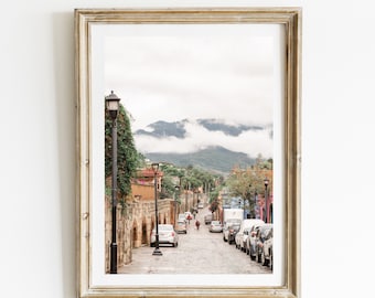 May include: Framed print of a cobblestone street in a town, lined with parked cars and buildings. A mountain range is visible in the background, partially obscured by clouds. The frame is gold-colored.