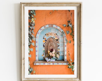 May include: A framed photograph of a religious shrine with a statue of the Virgin Mary, surrounded by blue and white mosaic tiles, and a bouquet of flowers in a purple and white patterned pot. The shrine is set against a bright orange wall with green ivy growing on either side.