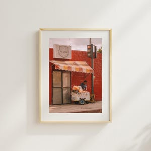 May include: A red brick building with a striped awning and a sign that reads "Pa' Llevar". A person is standing in front of a food cart selling fruit.