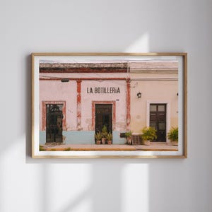 May include: Framed photograph of a building facade with three doorways. The building is painted in shades of pink, white, and beige, with the words "LA BOTILLERIA" above the center door. Potted plants are visible near the doors.
