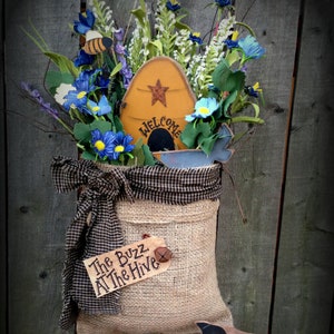 May include: A burlap bag with a black and white checkered ribbon, a wooden sign that says "Welcome", and a tag that says "The Buzz At The Hive". The bag is decorated with blue and white flowers and a bee. The bag is hanging on a wooden fence. The hashtag #K217 is written on a wooden surface below the bag.