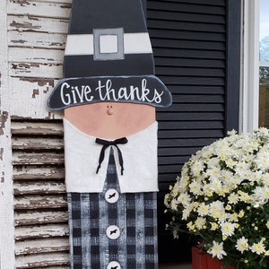 Può includere: Un ritaglio di legno di pellegrino con una camicia a quadri bianchi e neri, un colletto bianco e un cappello nero. Il ritaglio dice "Give thanks" sul cappello. Un ritaglio di legno di tacchino con un corpo marrone, piedi gialli e piume rosse e arancioni è in primo piano. Il tacchino ha un papillon a quadri bianchi e neri e un cartello che dice "Gobble". Ci sono zucche sullo sfondo.