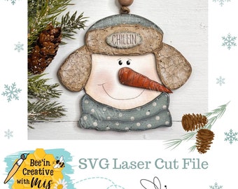May include: A wooden snowman ornament with a blue and white polka dot scarf, a brown hat with the word "Chillin'" on it, and a carrot nose. The ornament is hanging from a string of wooden beads.