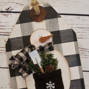 May include: A wooden snowman ornament with a black and white checked background. The snowman is wearing a black and white scarf and holding a small shovel and a potted evergreen plant. The ornament has a rustic, farmhouse style.