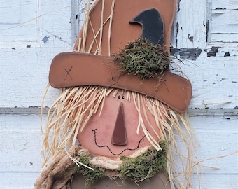 May include: A wooden scarecrow face with a brown hat, straw hair, and a burlap scarf. The face has a painted smile, nose, and eyes. The scarecrow is decorated with moss and straw.