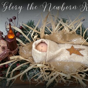 May include: A handmade nativity scene with a baby doll wrapped in a white blanket and a brown star. The baby doll is lying in a brown woven basket filled with straw. The scene is decorated with greenery, berries, and a lit candle.