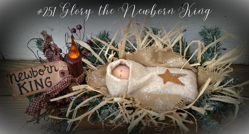 May include: A handmade nativity scene with a baby doll wrapped in a white blanket and a brown star. The baby doll is lying in a brown woven basket filled with straw. The scene is decorated with greenery, berries, and a lit candle.