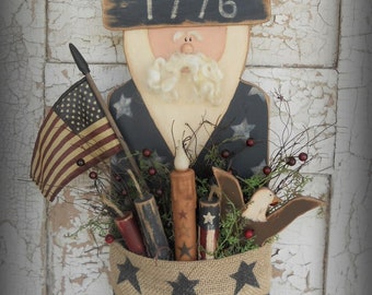 May include: A wooden patriotic figure with a red, white and blue top hat, a white beard, and a blue and white star-patterned shirt. The figure holds a burlap bag with a blue and white star-patterned ribbon and a tag that reads "America the beautiful".