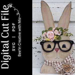 May include: A wooden bunny rabbit cutout with a flower crown, glasses, and a gingham bow tie. The bunny is painted in a light brown color and has a name tag that says "Hazel".