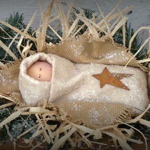 May include: A handmade nativity scene with a baby doll wrapped in white fabric and lying in a brown burlap manger. The manger is surrounded by straw and a small, rusty star.