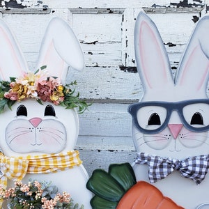 May include: Two white bunny decorations with painted faces. One bunny is wearing a yellow and white checkered scarf and a flower crown. The other bunny is wearing black and white checkered bow tie and glasses. The bunny with the glasses is holding a carrot.