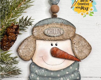 May include: A wooden snowman ornament with a blue and white polka dot scarf, a brown hat with ear flaps, and a carrot nose. The snowman's face is painted with a smile and black eyes. The ornament has the word "Chillin'" written on the hat.