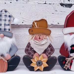 May include: Three wooden gnomes sitting side by side. The gnome on the left is wearing a blue and white plaid hat and holding a small orange pumpkin. The gnome in the middle is wearing a brown hat with straw and holding a yellow sunflower. The gnome on the right is wearing a red Santa hat and holding a small snowman.