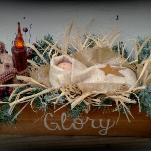May include: A rustic wooden manger with a baby doll inside, surrounded by straw, greenery, and a lit candle. The manger is decorated with a sign that reads "Newborn King" and the words "Glory" are painted on the front.