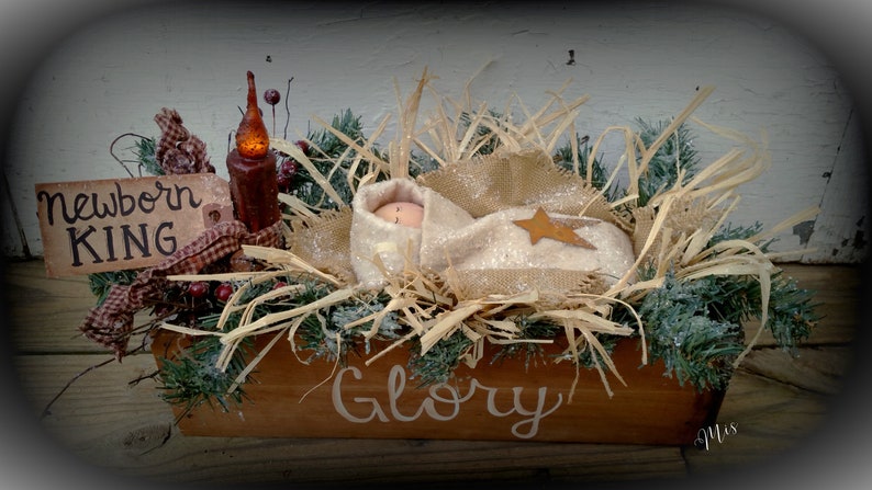 May include: A rustic wooden manger with a baby doll inside, surrounded by straw, greenery, and a lit candle. The manger is decorated with a sign that reads "Newborn King" and the words "Glory" are painted on the front.