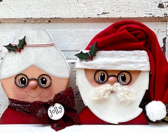 Puede incluir: Un par de adornos navideños de madera que representan a Santa Claus y a la señora Claus. Santa Claus lleva un gorro rojo con ribete blanco y una barba blanca. La señora Claus lleva una bufanda roja con estampado de cuadros y una diadema blanca. Ambos adornos tienen una expresión alegre y sostienen una rama de acebo.