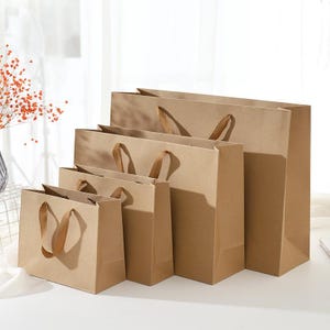 Könnte beinhalten: Ein Set aus vier rechteckigen Einkaufstüten aus braunem Papier in verschiedenen Größen, die in einer absteigenden Reihe vom kleinsten zum größten angeordnet sind. Jede Tüte hat einen flachen Boden, Seitenfalten und zwei passende Papiergriffe.