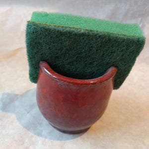 May include: A handmade, red ceramic sponge holder with a curved opening, holding a green and yellow kitchen sponge. The holder has a glossy finish and a rounded base. The sponge is rectangular and has a textured surface.