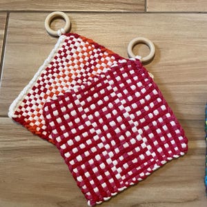 Könnte beinhalten: Zwei gewebte Topflappen mit rot-weiß karierten Mustern. Ein Topflappen hat einen Farbverlauf von Rot und Orange. Beide haben einen weißen Rand und einen Holzring zum Aufhängen.