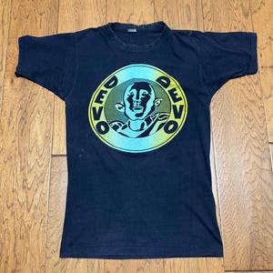May include: A navy blue t-shirt featuring a circular graphic with the word "DEVO" in yellow around a central image of a stylized face. The graphic has a teal and yellow gradient. The shirt is vintage and shows signs of wear.