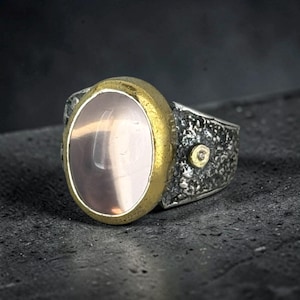 May include: A ring featuring a large, oval, pale pink cabochon stone set in a thick, textured gold-tone bezel. The band is made of dark, oxidized silver with a rough, cratered surface and a small, circular gold-tone accent on the side.