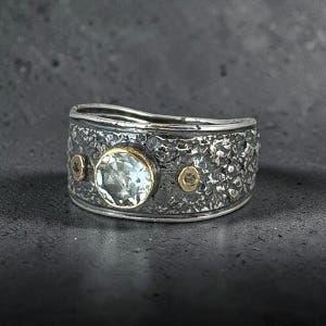 May include: A wide silver ring with a heavily textured, dark oxidized finish. The band features a central round, light green faceted gemstone set in a gold bezel, flanked by two smaller, circular gold accents, each containing a tiny embedded stone.