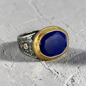 May include: A ring featuring a large, oval, faceted deep blue stone set in a thick, textured gold-tone bezel. The band is made of oxidized silver with a rough, cratered surface and includes a small, round, clear accent stone embedded on the side.