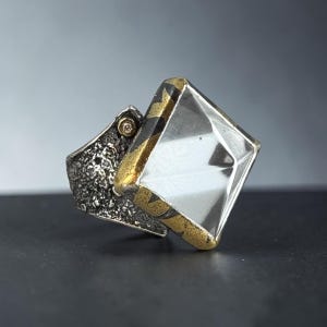 May include: A ring featuring a large, clear, square-cut faceted stone set in a gold-toned frame with a dark, textured, oxidized silver band. A small, singular, clear accent stone is embedded into the side of the textured silver metalwork.