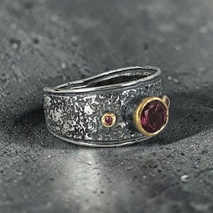 May include: A wide band ring featuring a dark, oxidized silver finish with a heavily textured surface. The ring is accented with a prominent round, deep pink gemstone set in a gold bezel, alongside two smaller, matching pink stones set in gold.